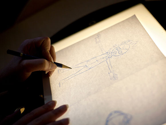 A close up photo of a hand holding a sheet of paper securely on top of a light box while the other hand draws with a pencil.