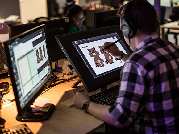 A person wearing a black and white plaid shirt and headphones leans over a tablet to create a digitally animated character.