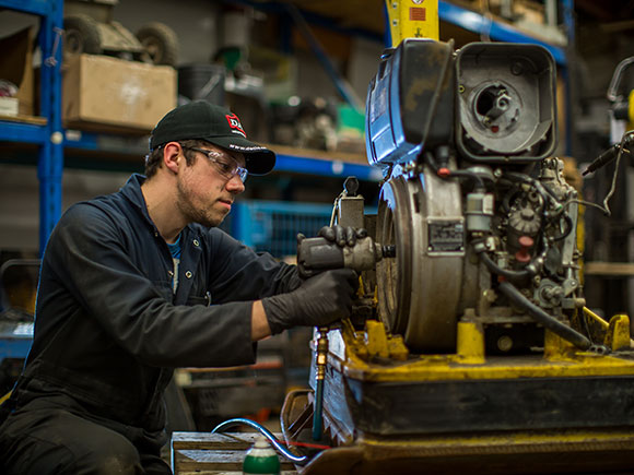 A man wearing a baseball hat, safety glasses and coveralls works on machinery in a workshop.