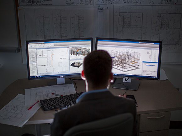 A man sits at a desk and looks at technical architecture drawings on two side-by-side computer monitors.