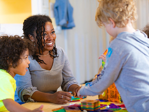 A woman sits at a small table and smiles at two toddlers who are playing with her.