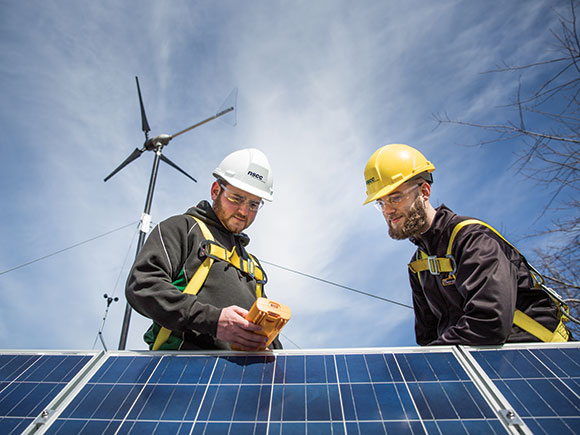 Two men in hard hats, safety goggles and safety harnesses hold a piece of equipment while standing near solar panels.