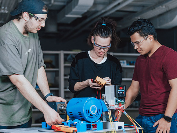 Three students in safety glasses learn about equipment in a campus workshop.