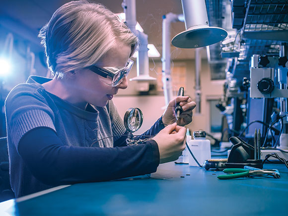 A woman sits at a desk and works on electronic equipment in a campus learning lab setting.