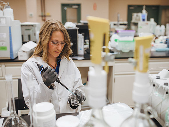 A woman wearing safety glasses, a white lab coat and gloves tests water in a laboratory setting.