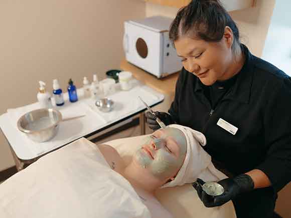 A smiling student practices giving a facial to someone in a spa setting.