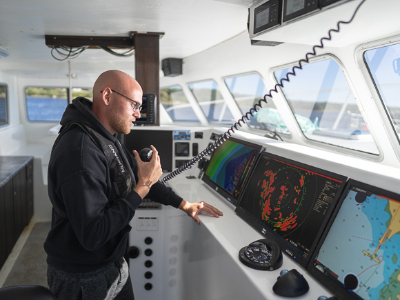 A person wearing a hoodie and a personal floatation device navigates a fishing vessel while using a radio.