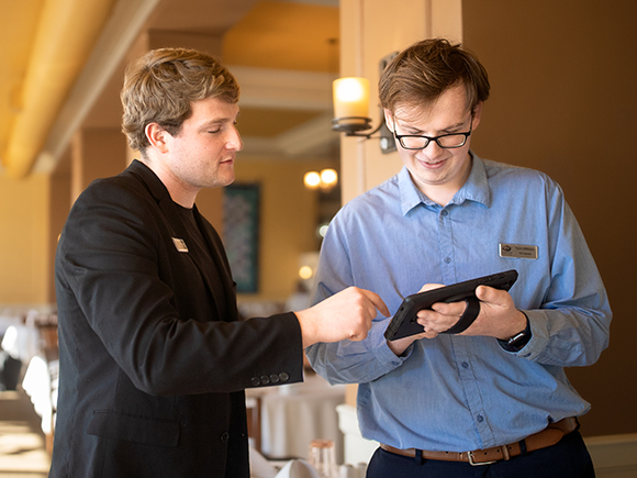Two people wearing professional clothing and name tags stand in a restaurant setting. One holds a tablet while other points t