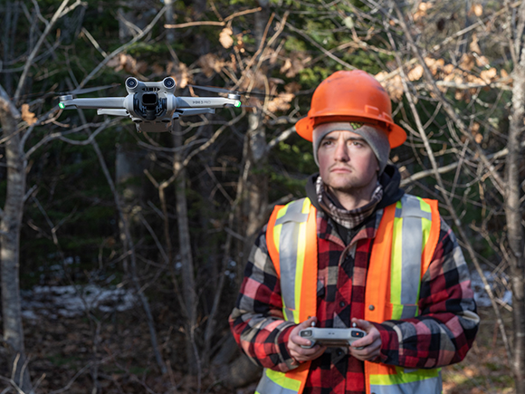 A man in a hardhat and safety vest operates a drone in a forest.
