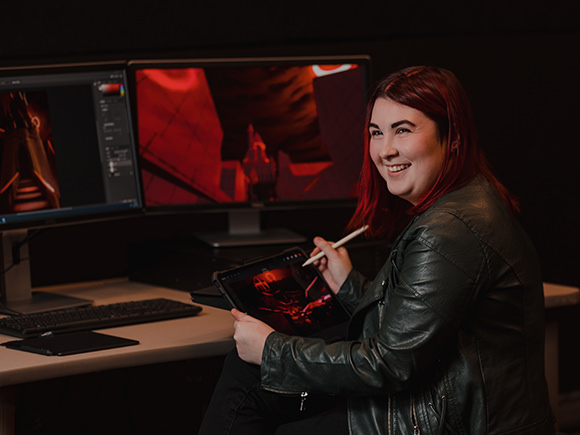 A student wearing a leather jacket sits in front of multiple computer monitors and designs an element of a game.