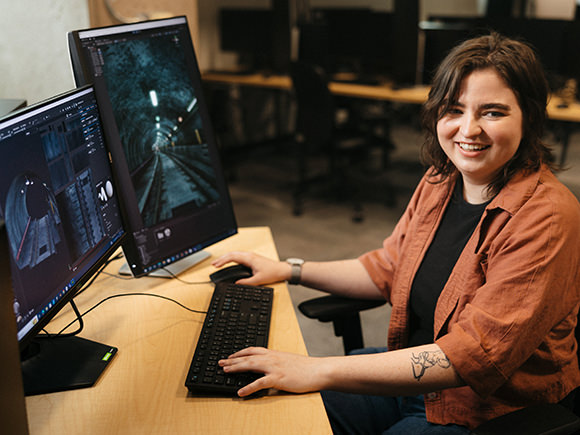 A smiling student in a computer lab sits at her computer with two large screens working on her interactive art.