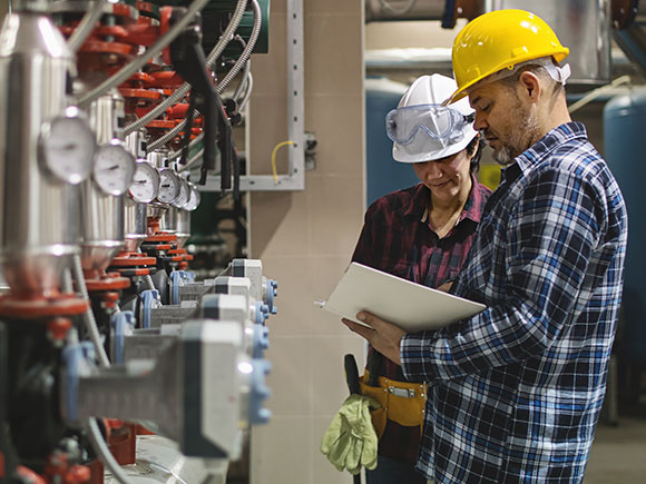 Two people in hard hats consult a piece of paper while standing on front of a row a propane tanks.