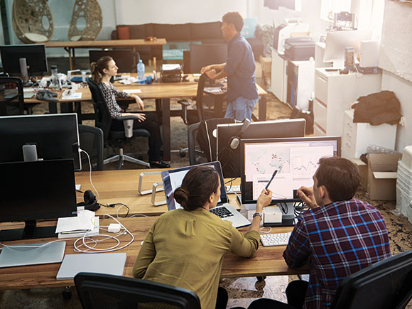 A man and woman in the geospatial analytics program look at a computer screen while two other people chat in the background.