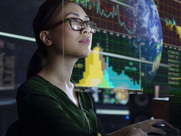 A woman wearing glasses works at a computer station and examines GIS data.