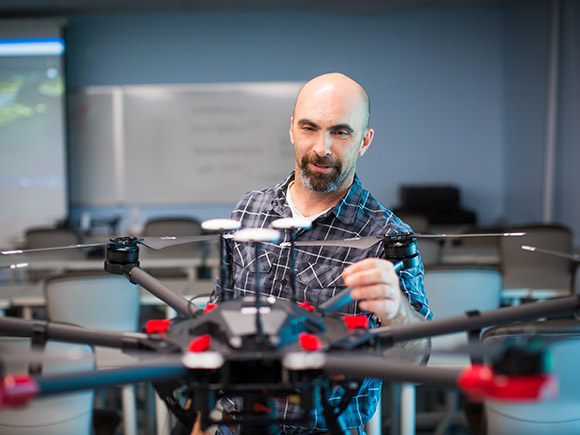 A man wearing a plaid, button-down shirt examines a drone in a classroom setting.