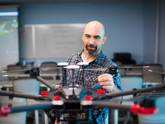 A man wearing a plaid, button-down shirt examines a drone in a GIS Remote Sensing classroom.