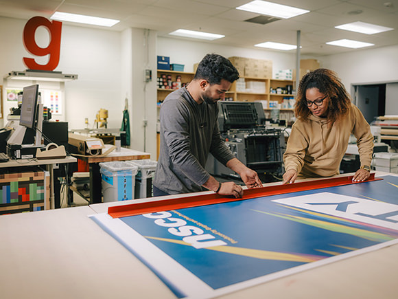 A man and woman examine a large banner laid out on a table in a print shop.