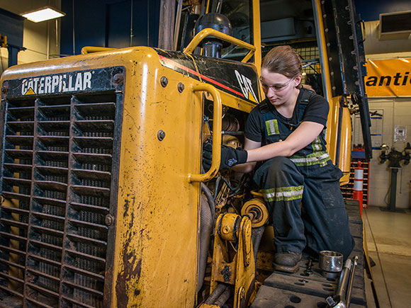 A woman in overalls works on a yellow tractor.