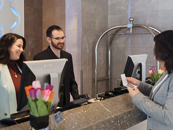Two people stand behind the dront desk of a hotel lobby and smile at a guest who has just been handed a room key.