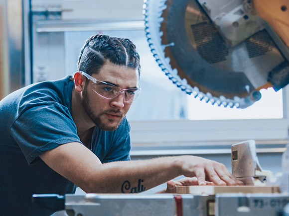 A man wearing safety goggles uses a chop saw in a construction workshop.
