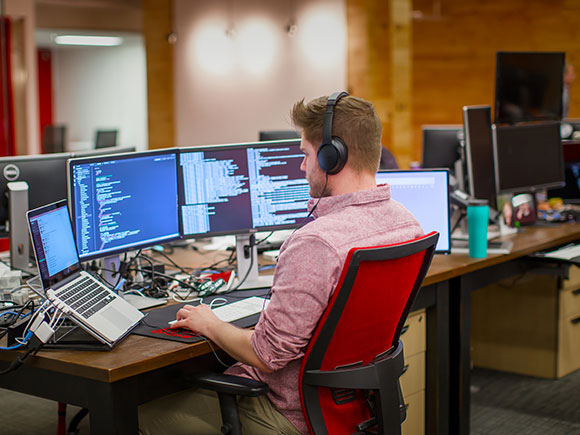 A man wearing headphones sits at a desk with five computer screens in front of him.