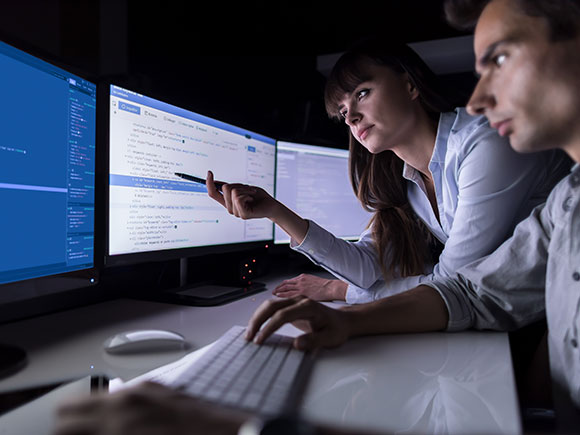 A man and woman gather around three computer screens; the woman points to one of the screens with her pen.