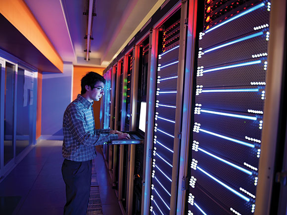 A man works on a computer while standing in a colourful computer server room.