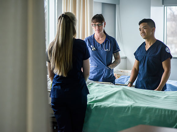 Two female nurses and one male nurse talking with a patient in their bed.