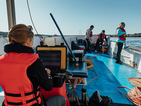 Four people, all wearing life jackets, work on the deck of a small boat; one woman has a laptop in front of her.