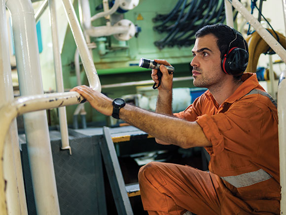 A man wearing ear protection and orange coveralls examines a ship's engine room with a flashlight.