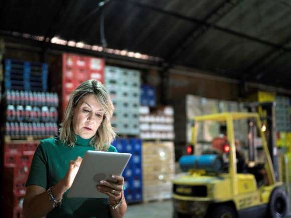 A woman with a tablet works while a forklift operates in the background.
