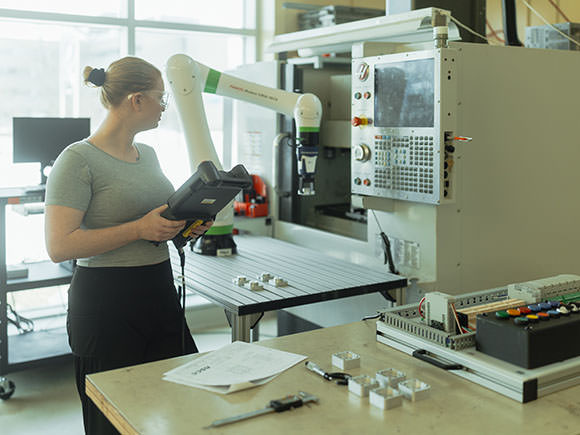 A woman in protective goggles using her tablet as reference, works on a robotic arm machine.