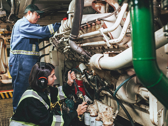 Two people, one wearing ear protection, work in a ship's engine room.