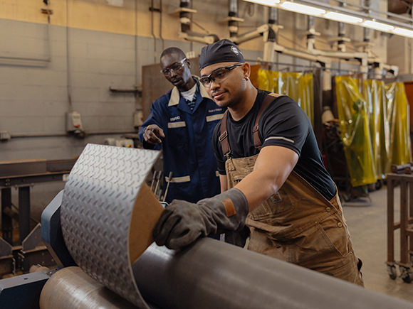 A student wearing safety gear works on a piece of metal in a workshop setting with a supervisor looking at what's being done.