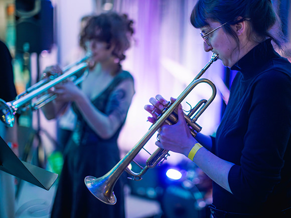Two women are seen on stage playing their trumpets at an event.