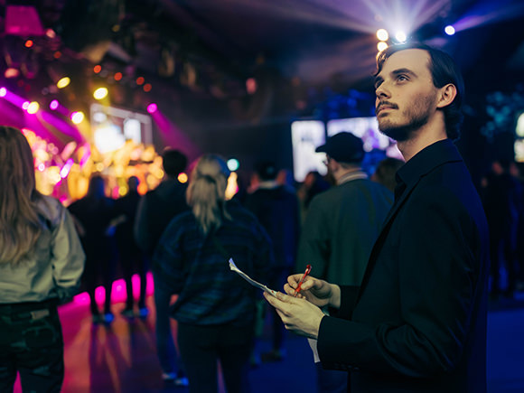 A man stands in a busy, lit up space with a clipboard in hand.