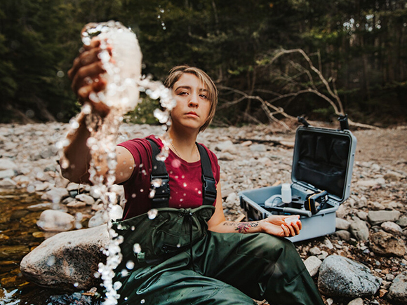 Kieran, a Natural Resources Environmental Technology program grad, sits on a rocky shoreline and fills a container with water