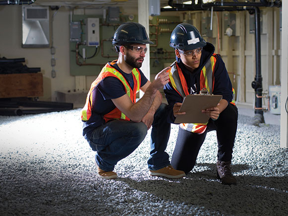 Two men in safety vests, goggles and hard hats consult a clipboard while kneeling in an unfinished basement or utility room.