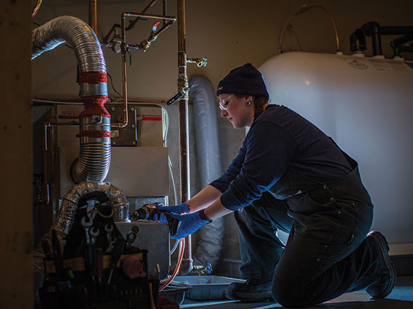 A woman in overalls kneels down to adjust a piece of equipment.