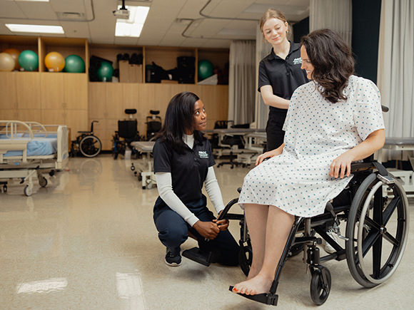 Two students wearing black shirt uniforms in a physiotherapy setting work with a patient sitting in a wheelchair, wearing a h