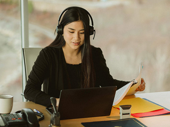 A woman with long, dark hair wearing headphones and sitting at a desk is seen working on a laptop while also holding document