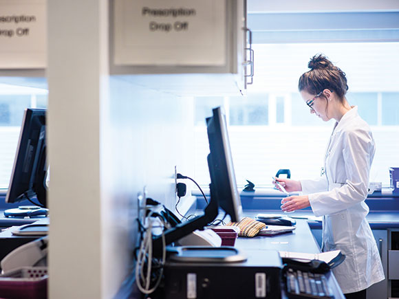 A woman in a lab coat works behind a prescription drop-off counter.