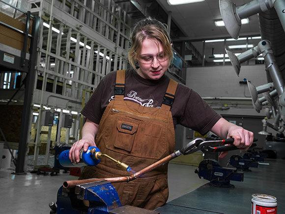A woman in brown overalls and safety glasses uses a blow torch on a piece of pipe.