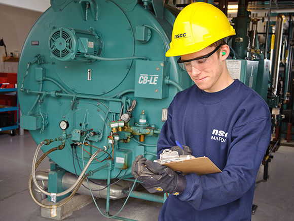 A man in a blue NSCC shirt and yellow NSCC hard hat stands in front of a large piece of equipment and writes on a clipboard.