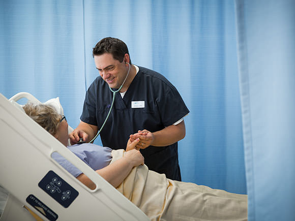 A nurse wearing scrubs smiles while standing near a patient's bed. He holds one of the patient's hands in his and uses a stet