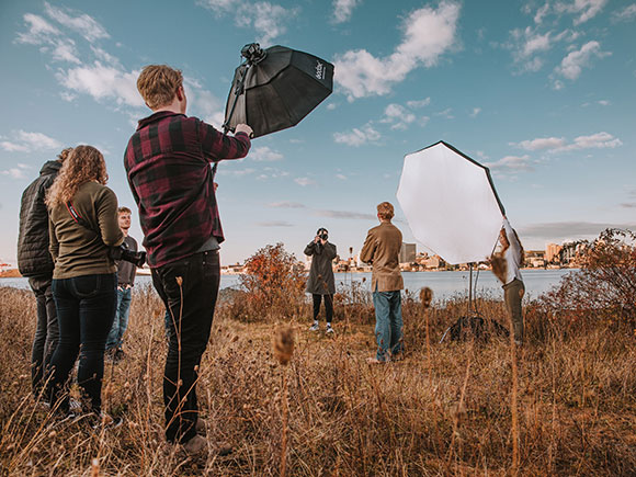 Students stand in a field for a photoshoot; one is posing, another is taking a photo and the rest are assisting or observing.