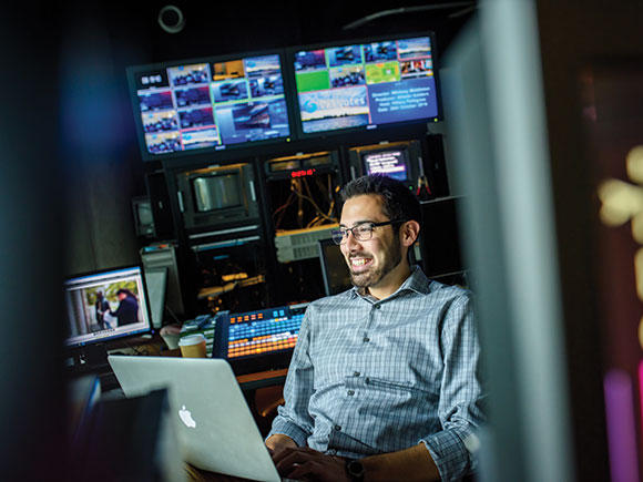 A man sits in a production studio; he's smiling and his laptop in on the desk in front of him.