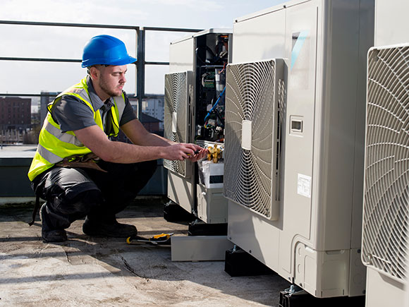 A man in a yellow safety vest and blue hard hat kneels beside an air conditioning unit.