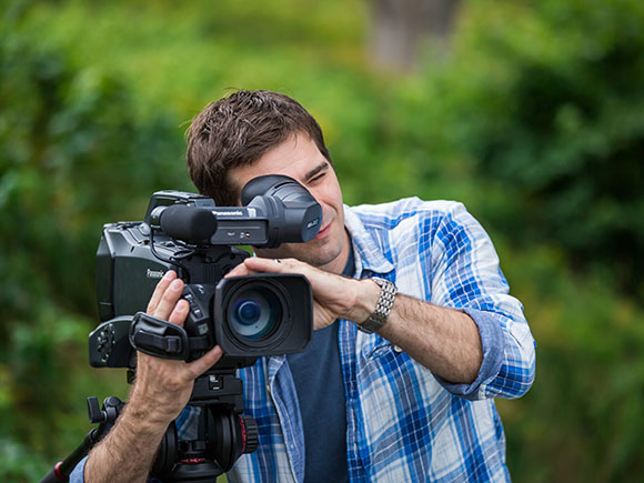 A man in a blue and white plaid shirt looks through a TV camera.