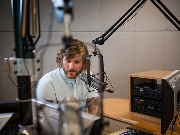 A man sits in front of a mic while seated in a radio studio.